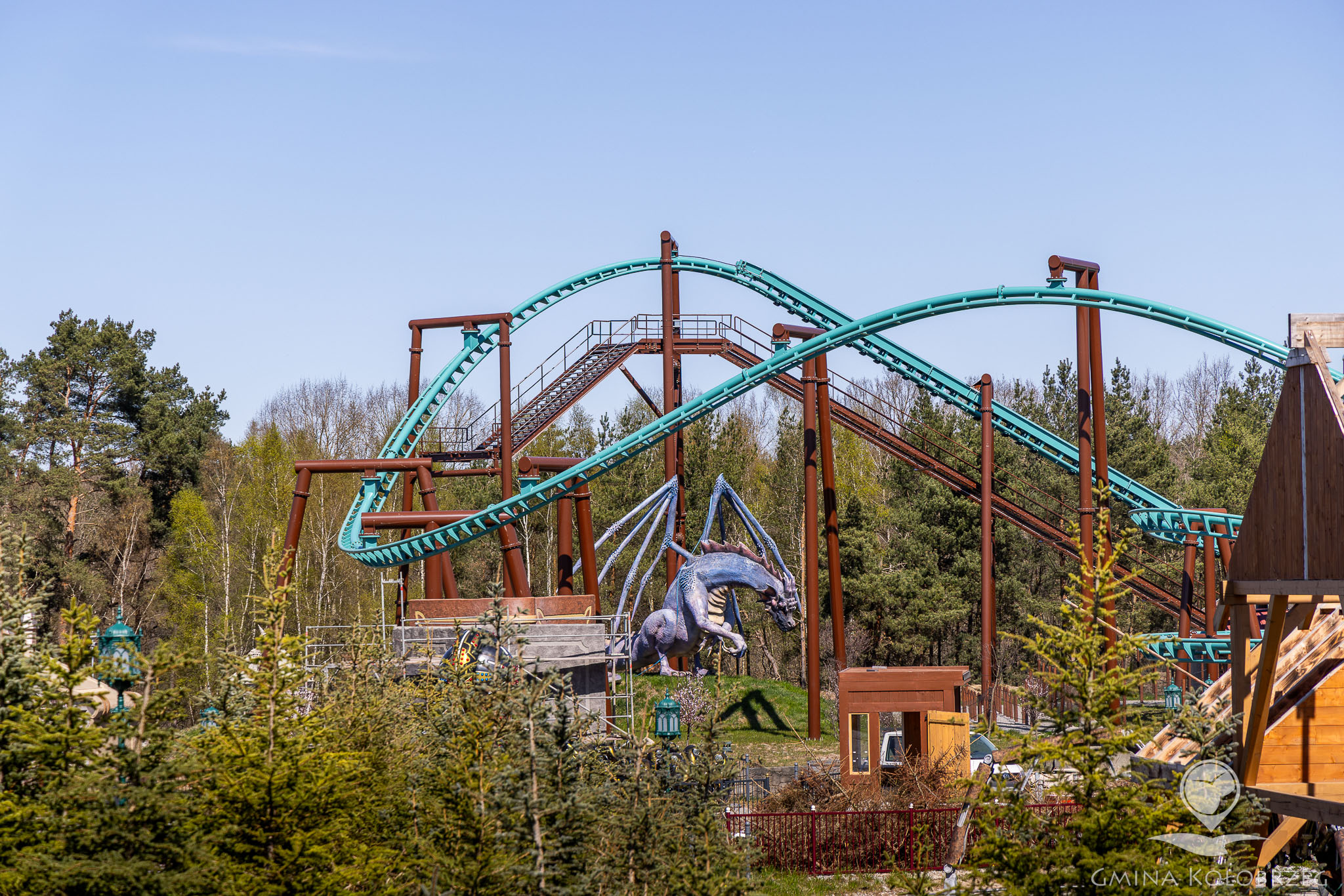 Na zdjęciu widać fragment parku rozrywki z kolejką g&oacute;rską o turkusowych torach oraz konstrukcją wsporczą. W centralnej części znajduje się dekoracja w formie smoka, co sugeruje tematyczną atrakcję. Otoczenie stanowią drzewa i elementy infrastruktury parku.