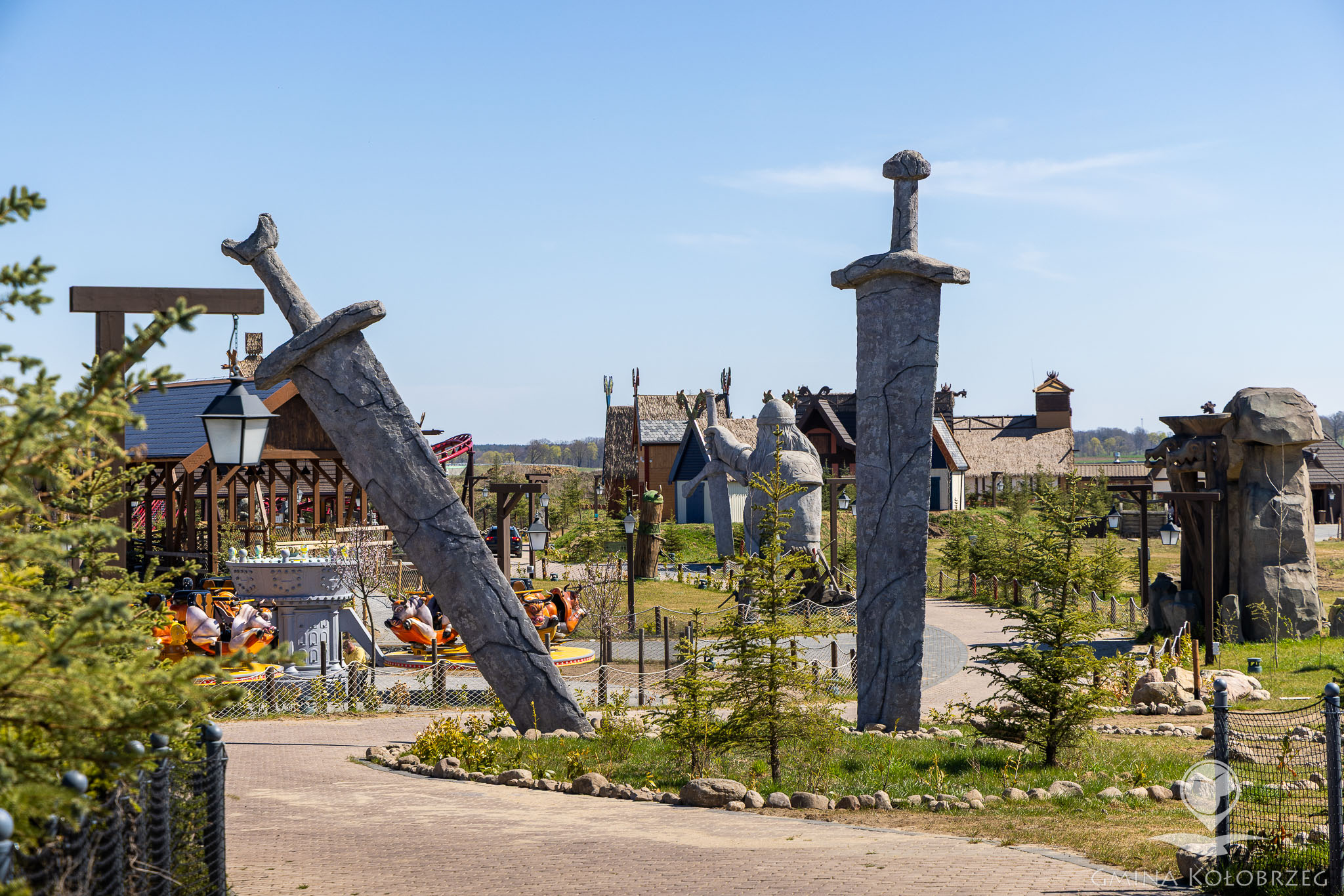 Na zdjęciu widać teren parku rozrywki stylizowanego na klimaty historyczne lub fantastyczne. Na pierwszym planie znajdują się duże dekoracyjne miecze wbite w ziemię, a w tle widoczne są atrakcje, zabudowania tematyczne oraz karuzele. Całość otoczona jest zielenią i ścieżkami spacerowymi.